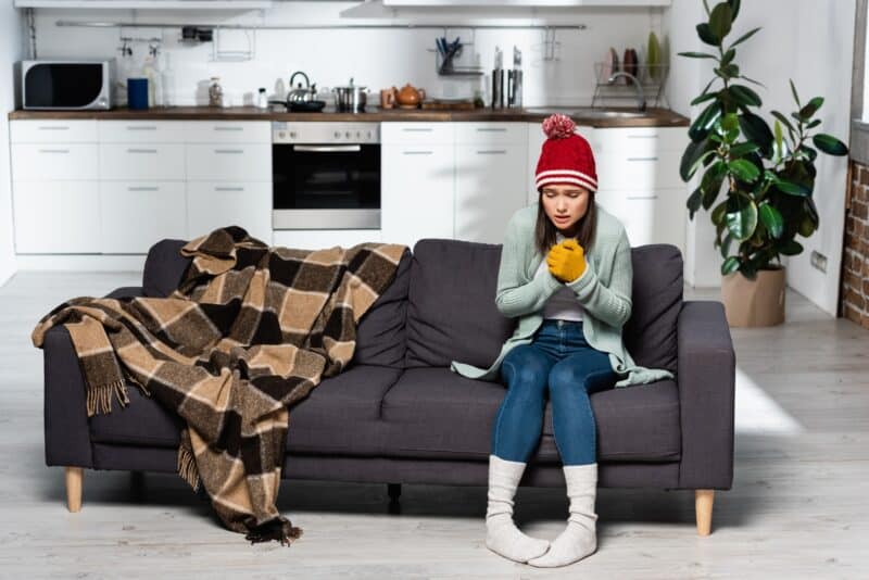 young woman sitting on her brown couch showing signs of being cold while sitting next to a blanket and wearing long socks and a beanie and gloves.